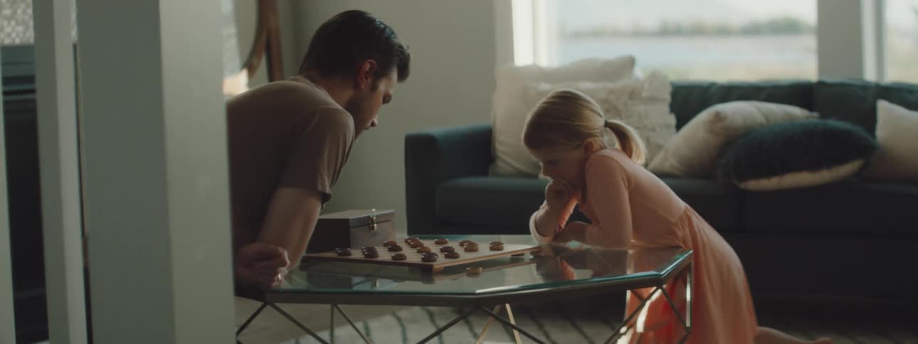 A father plays a game of checkers with his young daughter who hops up excitedly