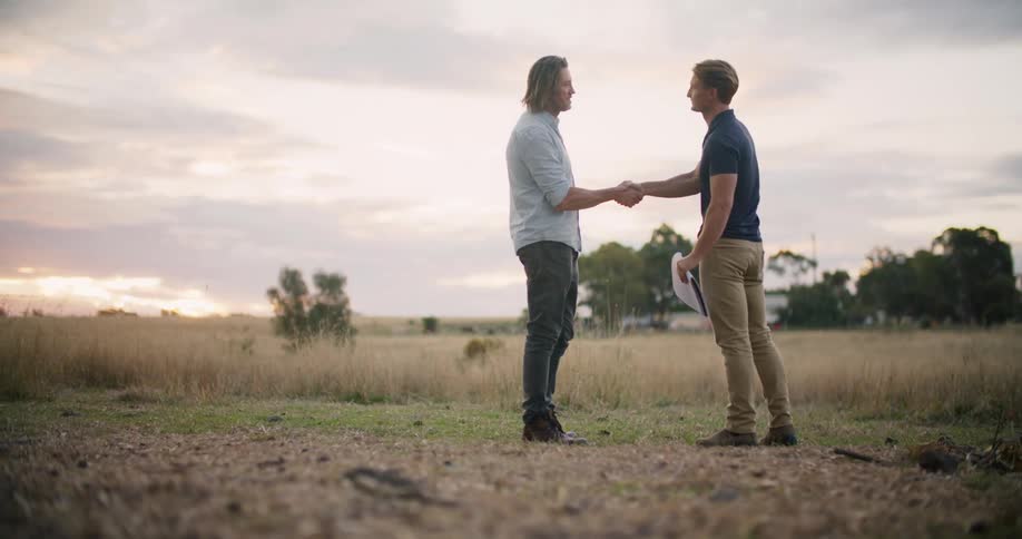 Two men are standing in a rural area and shaking hands at dusk