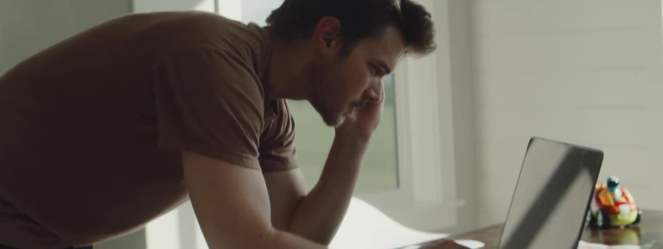 A man talks on the phone while leaning down and using a laptop at home