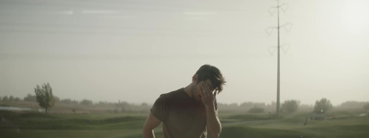 A man rubs his eyes and face as he stands near a rural field on a windy day