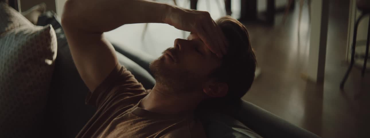 A man sitting on a couch at home leans his head back and rubs his temple