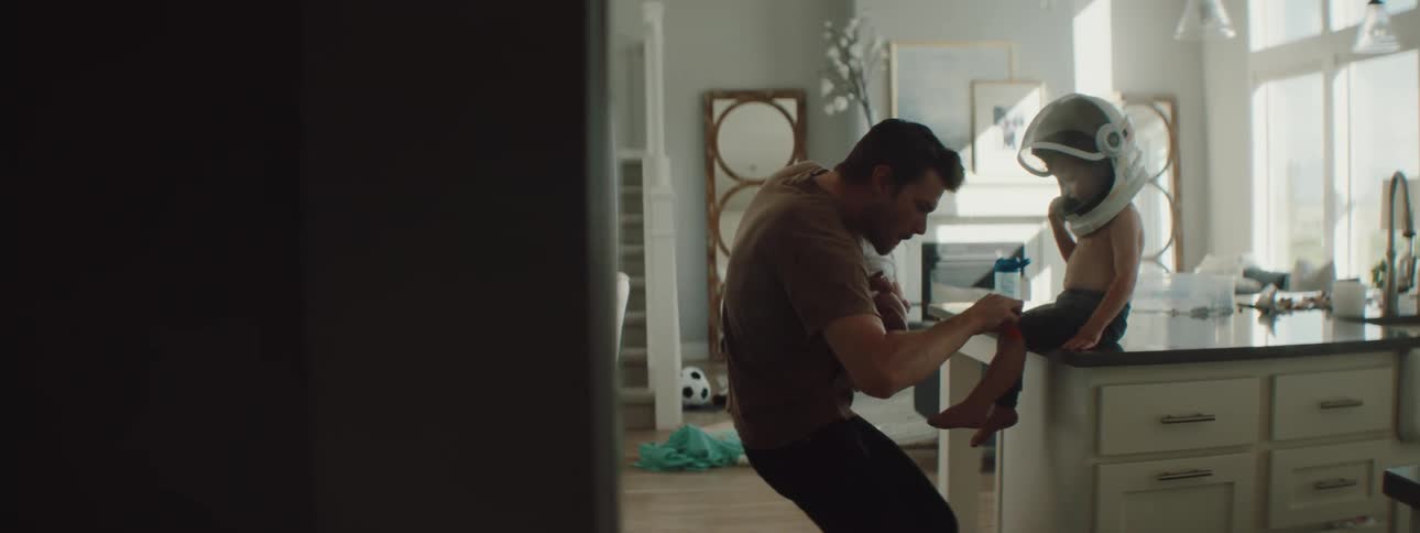 A man holding a young baby in one arm puts a bandaid on a young boys knee and picks him off the counter