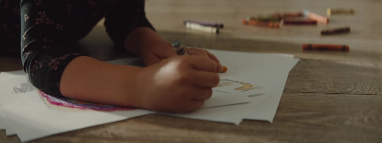 A girl laying on the floor coloring with crayons with chairs and a wall in the background
