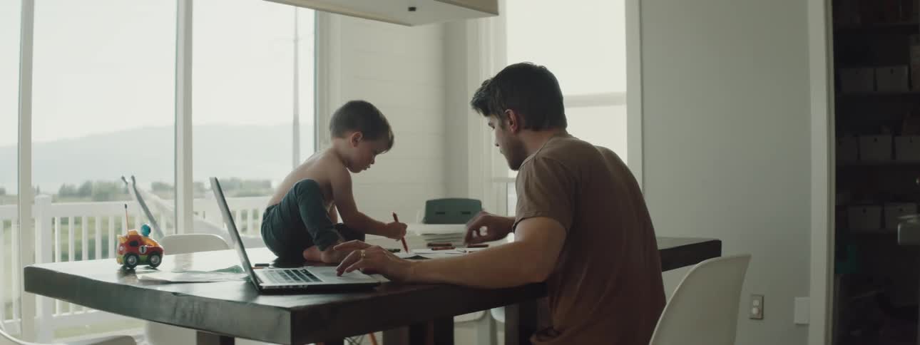A man works on his laptop at home as his son sits on the table and draws