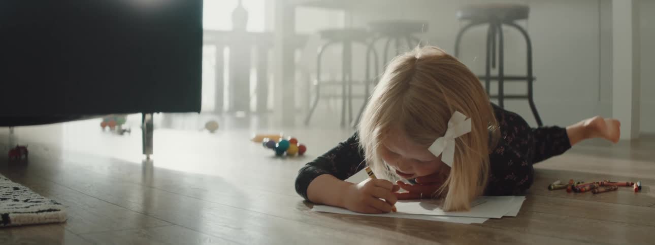 Little girl lays on the floor on her tummy and colors with crayons on white paper