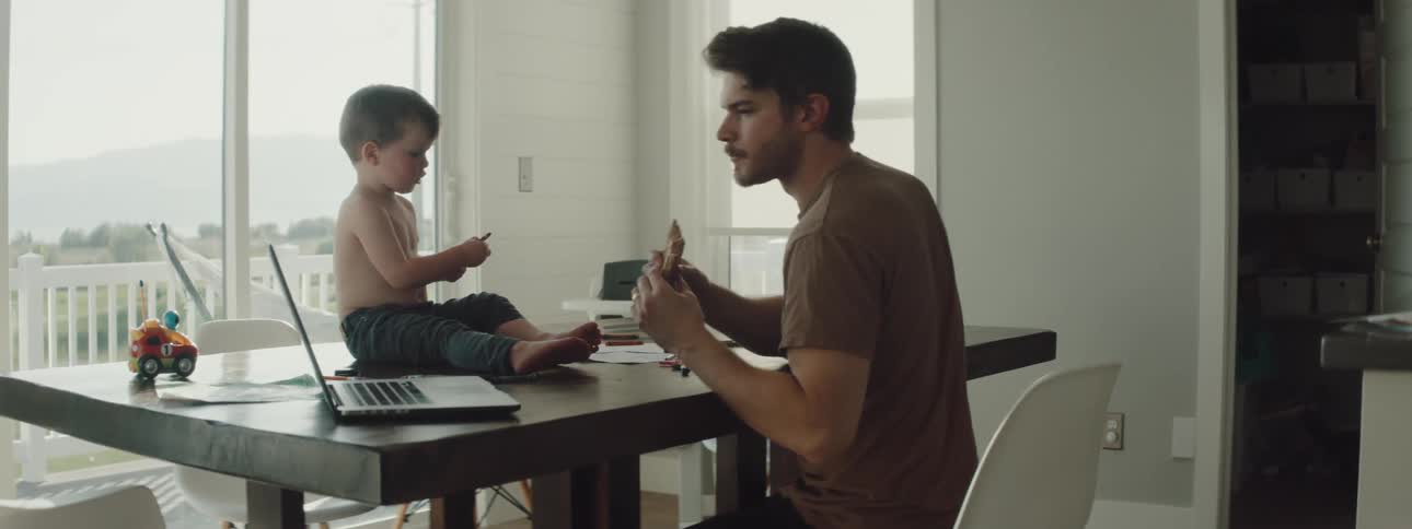 A man sitting on a chair at a table typing on a laptop with a boy sitting on the table