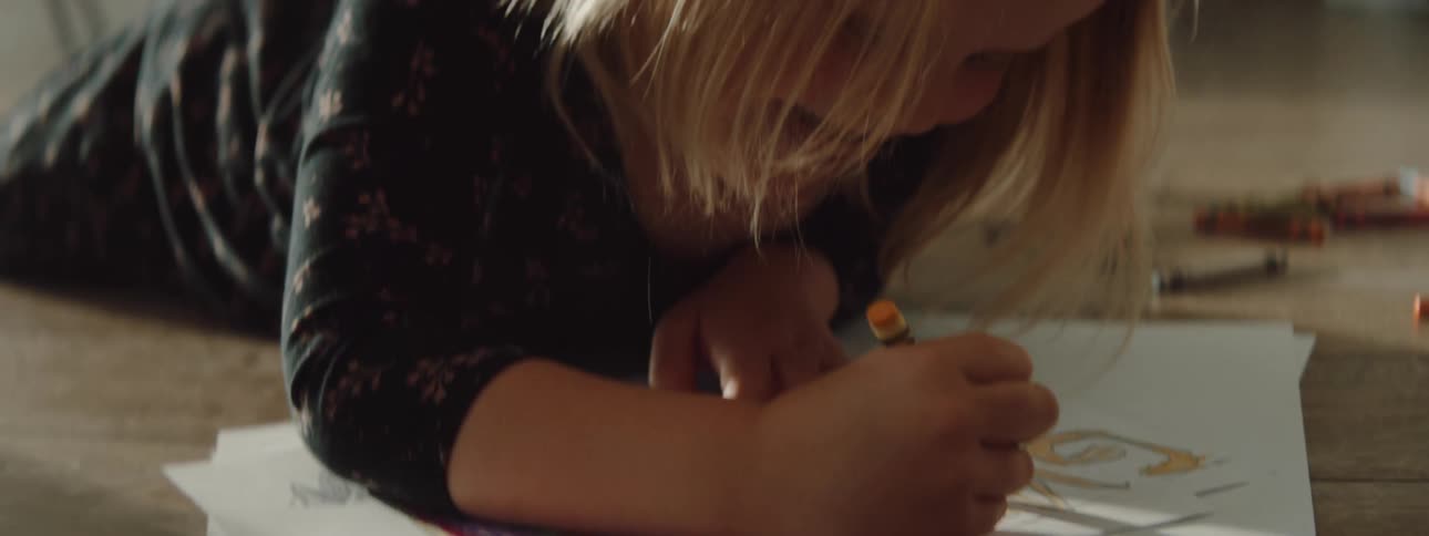 A young girl uses a crayon to color on a sheet of paper as she lies on the floor indoors during the day