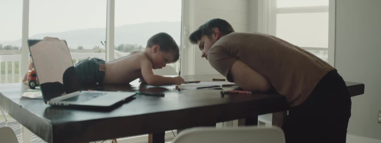 A man leans onto his kitchen table while watching his toddler son draw