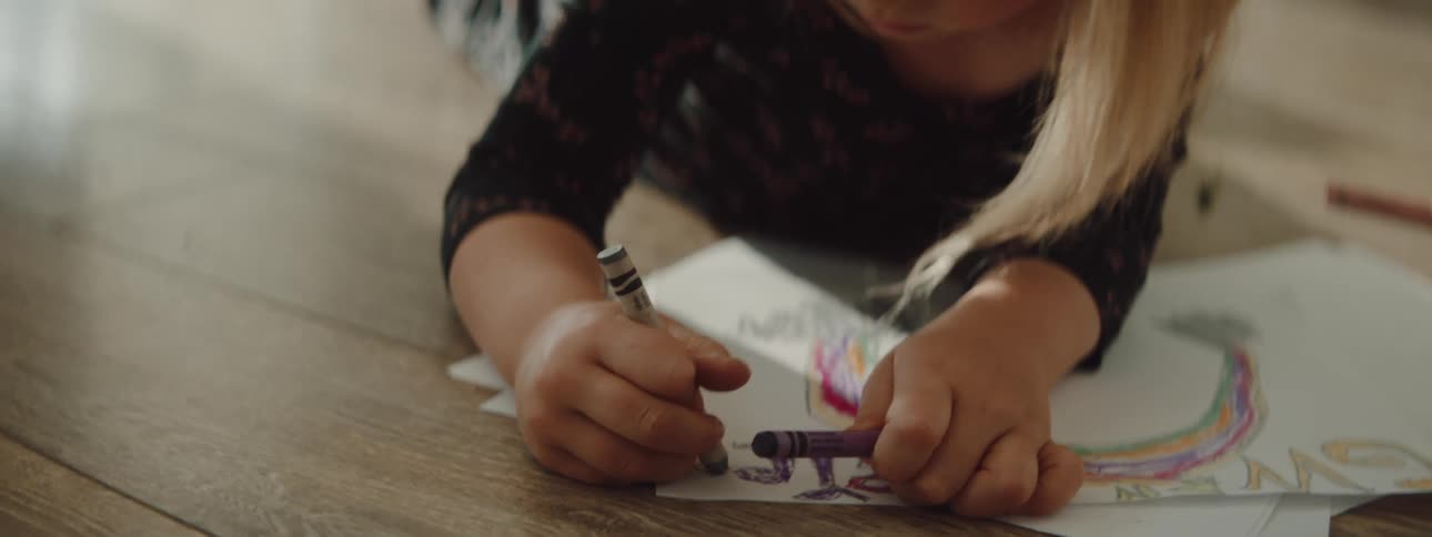 A girl laying on the floor coloring with crayons with chairs and a wall in the background