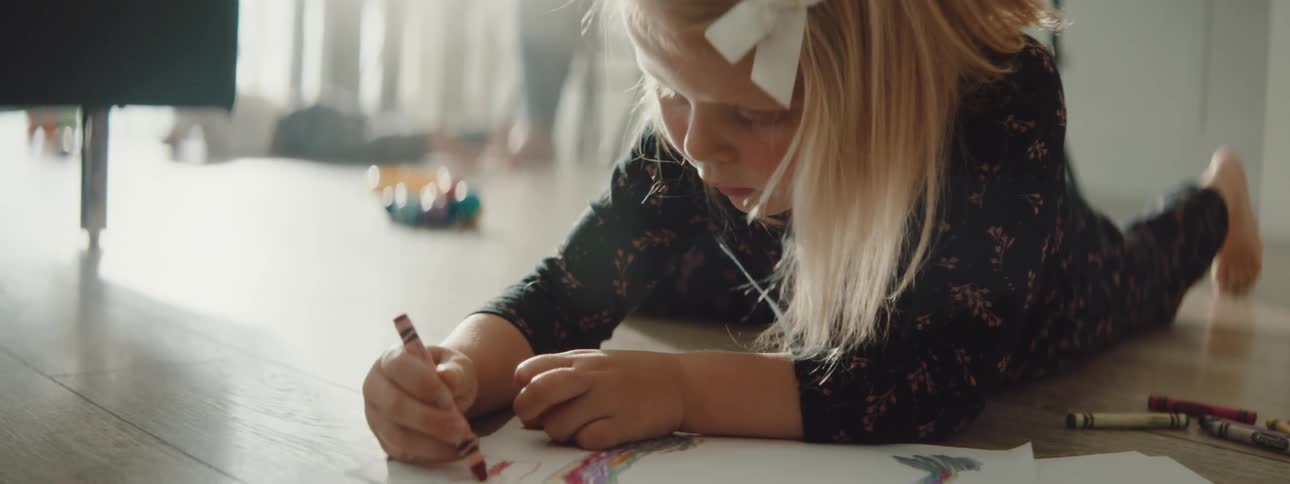 A girl in a room laying on the floor coloring with crayons on a paper