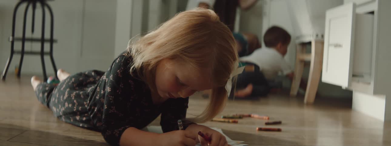 Mother works near son while daughter lies on stomach on floor using crayons