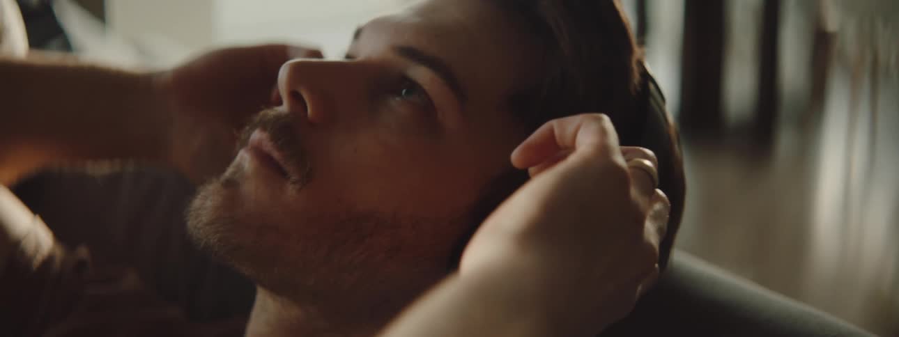A man adjusts his over the ear headphones as he sits with his head leaned back on his couch