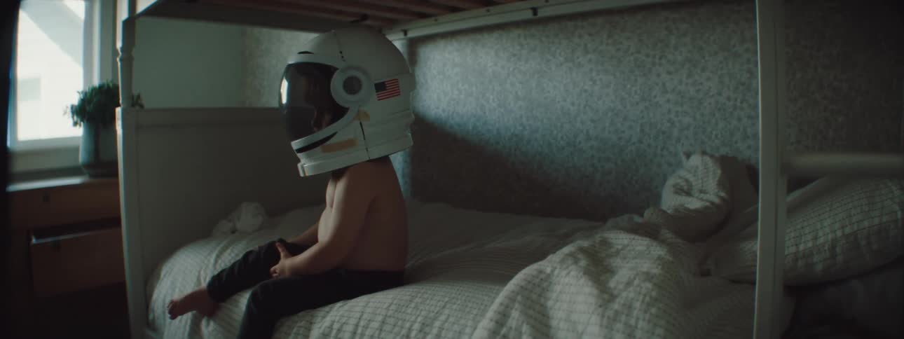 A little boy sitting on a bunk bed wearing a large astronaut helmet