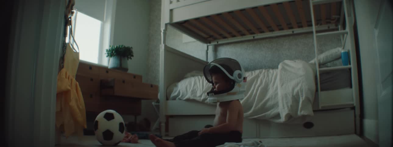 A young boy sits on the floor of his bedroom wearing an astronaut helmet