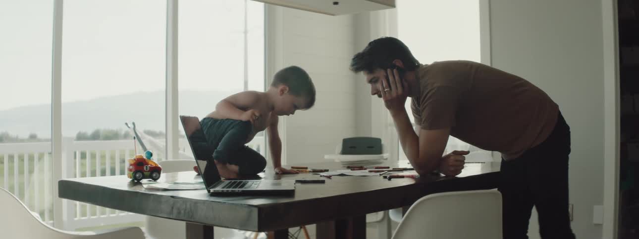 Father leans over table near laptop talking on mobile phone while son sitting on tabletop colors