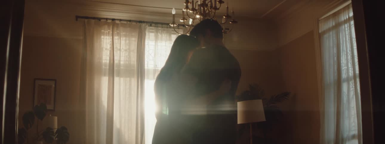 Couple holding each other and rocking gently standing in front of a flare of sunlight coming in from a window inside of a living room