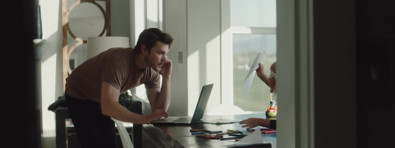 A father talking on a phone while standing and scrolling on a laptop computer on a table while his daughter shows him a piece of paper sitting at a dining room table