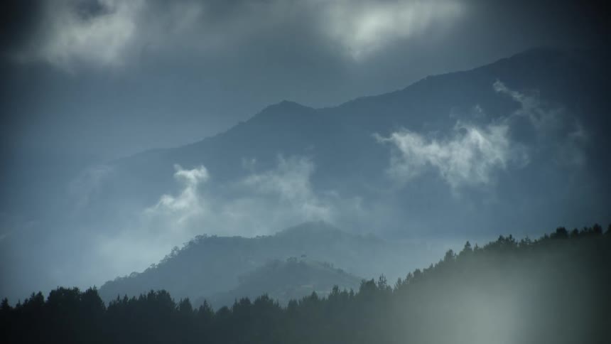 A tall mountain range towers above a sprawling forest as clouds drift past