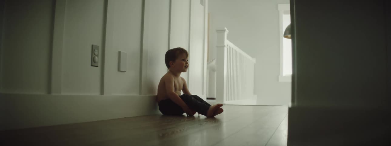 A boy sits on a wooden floor leaning against a wall in a hallway at home looking up and smiling