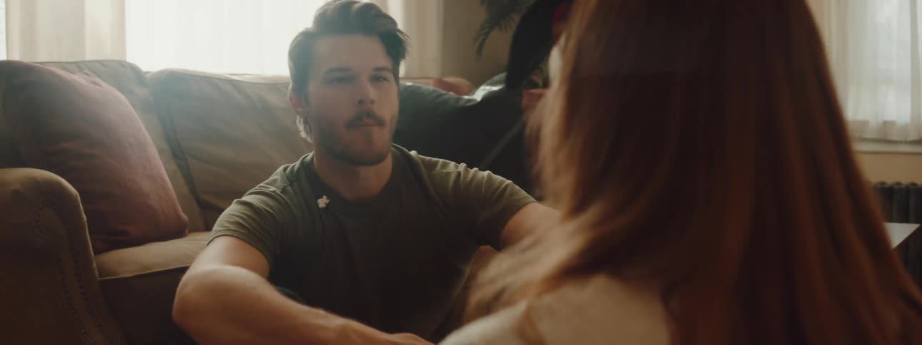 A man and woman sit on the floor in a house and the woman throws popcorn in the mans mouth