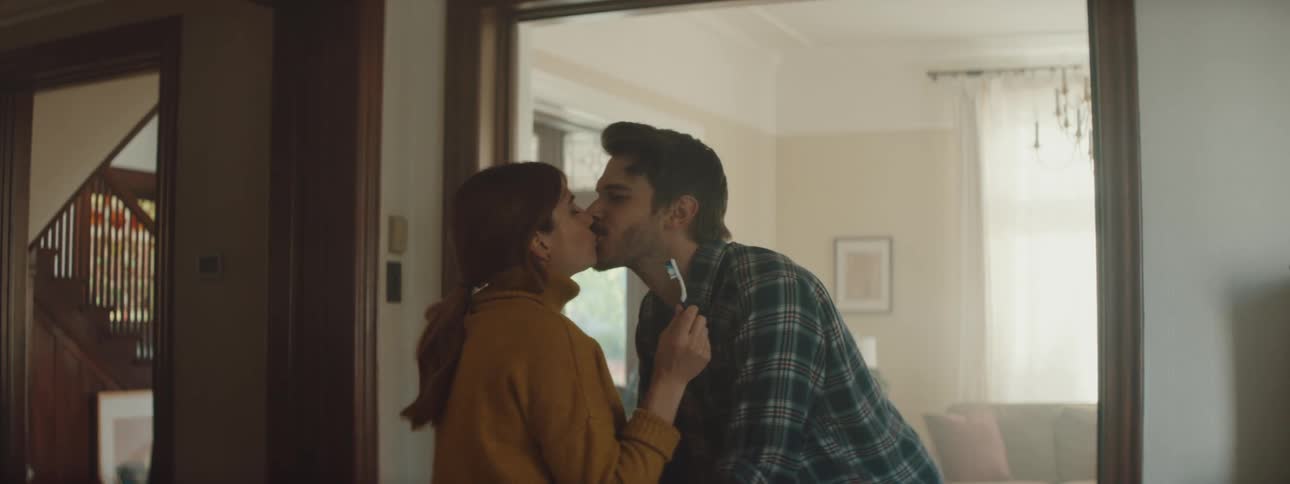 A woman laughs as her husband interrupts her brushing her teeth while standing near their living room