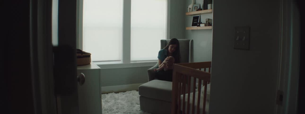 A mother holding her baby gets up from a rocking chair next to a window in a nursery and walks over to a dresser