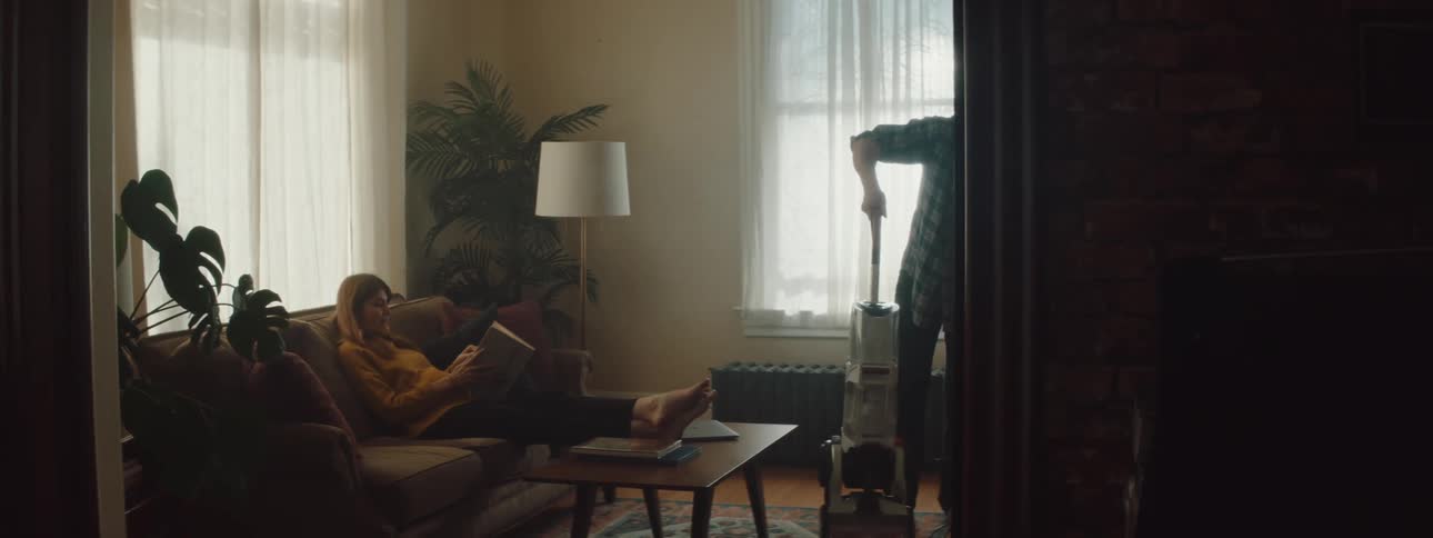 Man vacuuming the rug on the living room while his wife sits on the couch
