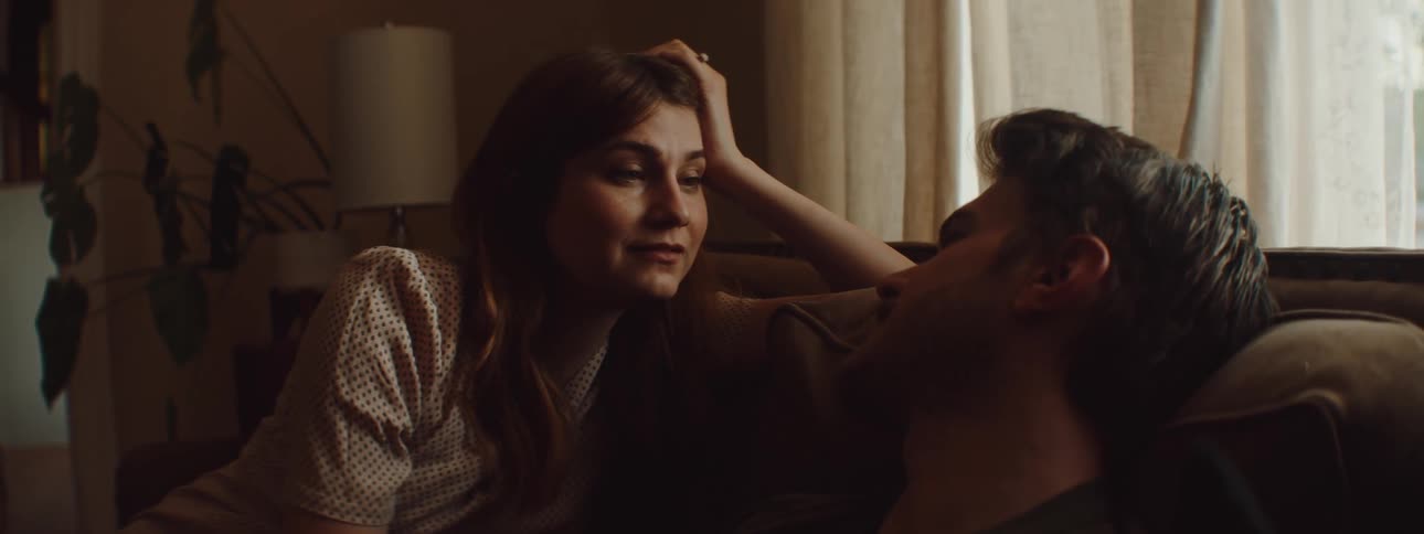 A man and a woman sitting on a couch as a woman strokes the mans hair with a wall and window in the background