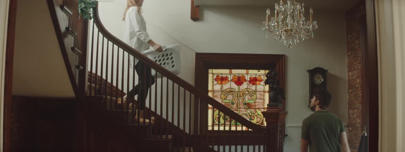 A man walks up the stairs in his home and kisses his wife who is carrying a basket of laundry down