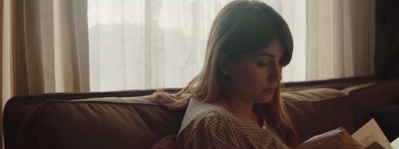 Woman with brown hair and a blouse reads a book in the living room at home
