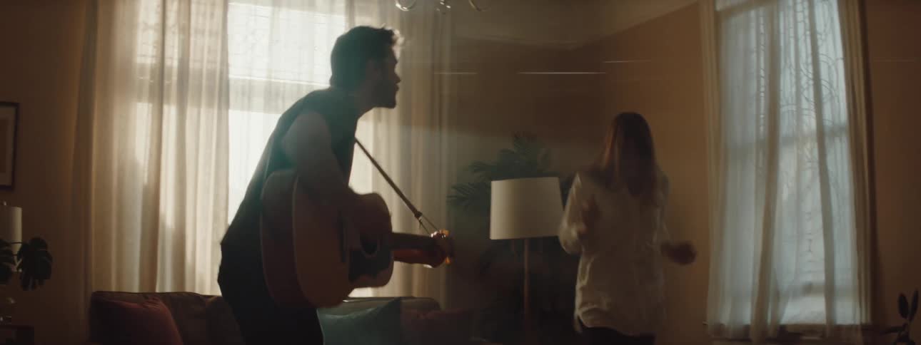 A man and woman sit on a couch together than he gets up and plays the guitar while she gets up to dance