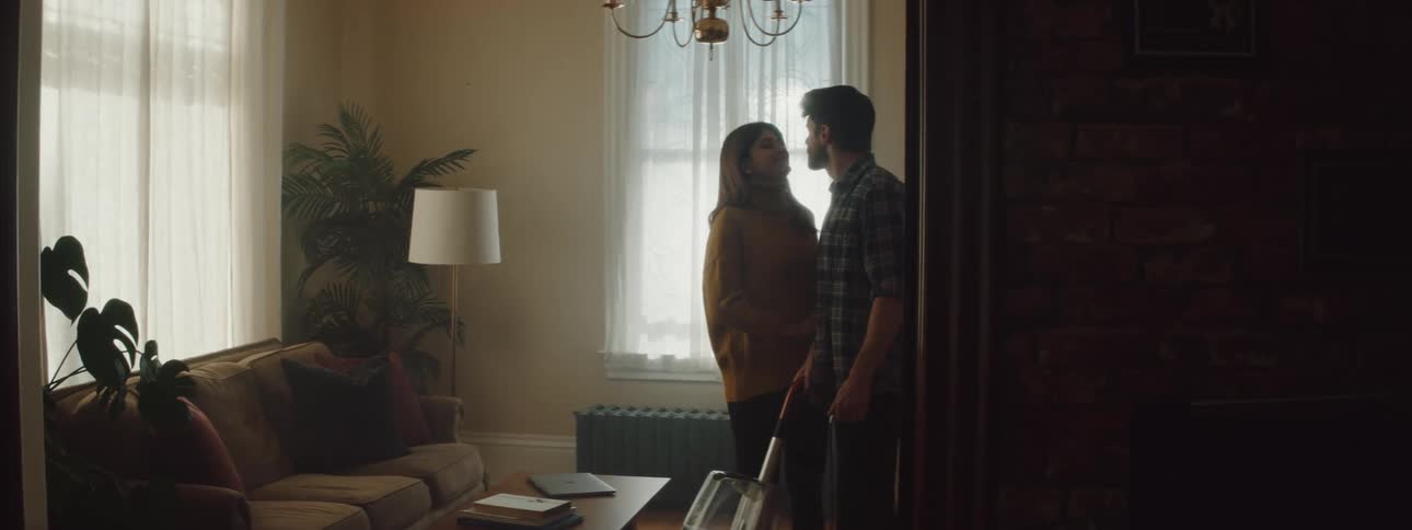 A man vacuuming the floor in a room as a woman adjusts cushions on a couch then kisses him
