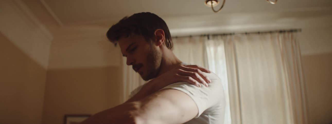 Man standing in room stretches arm and shoulder winces and then rubs shoulder