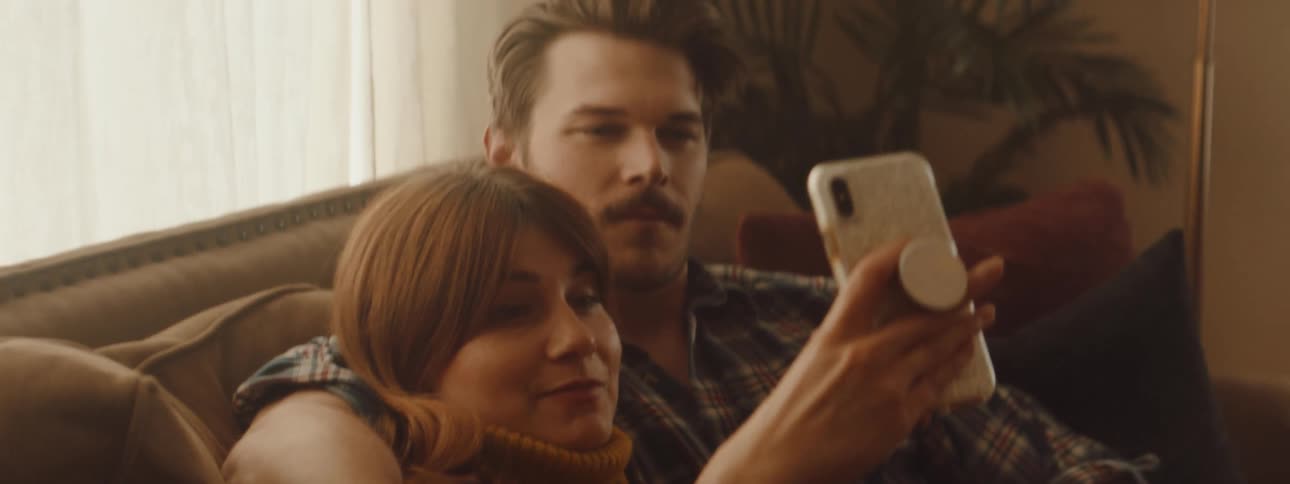Man with a mustache and a shirt holds woman with brown hair and a yellow turtleneck sweater using a smartphone in the living room at home