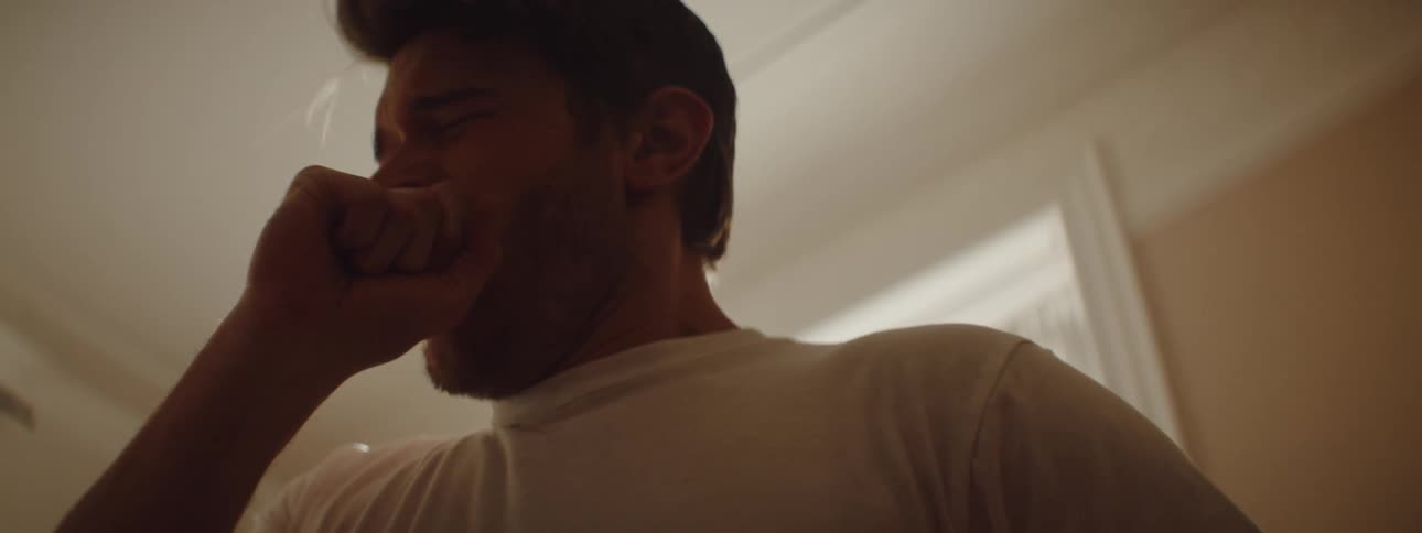 Man stands near a window and rubs his eyes with his hand and then yawns into his fist