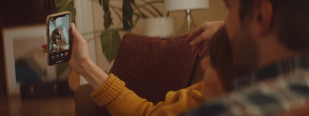 A couple lie on the couch together while talking to a toddler on a video call on their smartphone