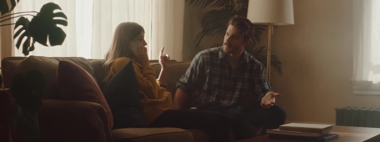 A man looks confused at the woman sitting beside him on the couch talking on the phone