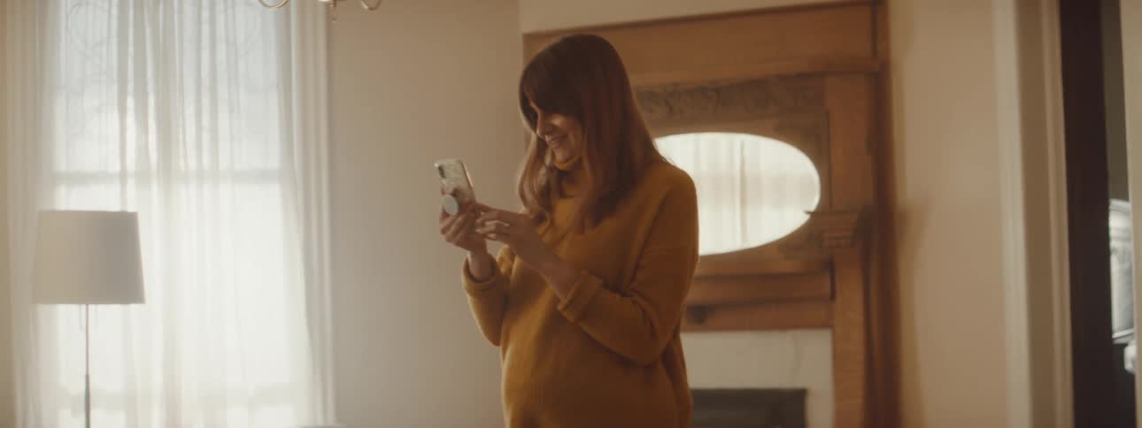Pregnant woman with brown hair and a yellow sweater makes a video call in the living room at home