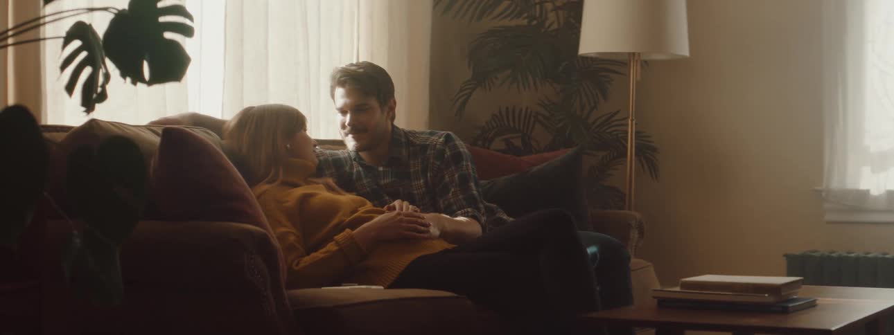 A man rubs the stomach of a pregnant woman resting next to him on a sofa in the living room of a house during the day