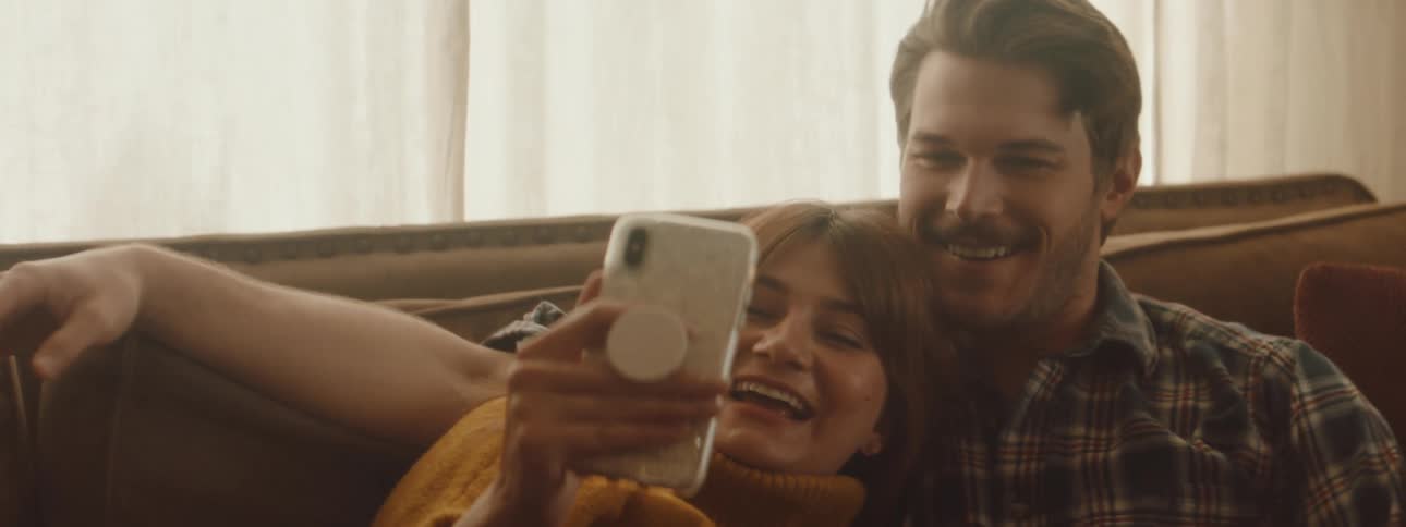 A man and a woman sitting on a couch looking at a smartphone with a window and blinds in the background