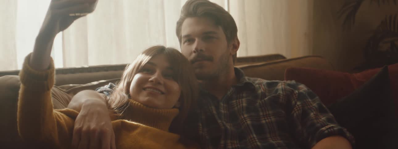 A man and a woman sitting on a couch taking a picture with a smart phone with a wall and window in the background