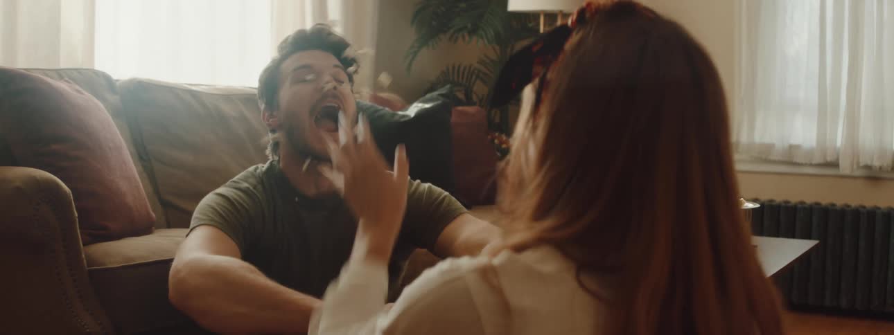 A man and woman sit on the floor and the woman throws popcorn at a mans face