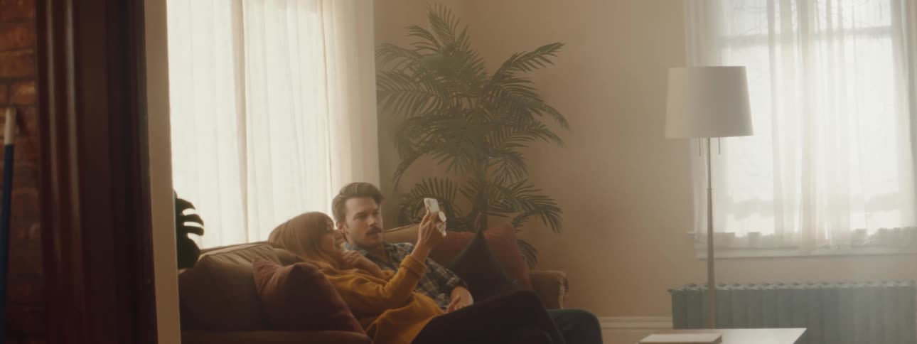 Young pregnant couple sits in a couch in the living room looking at the phone