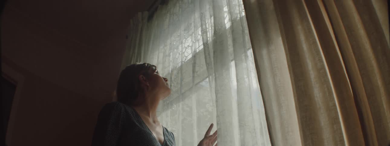 A woman in a room looking out a window with a curtain with a wall in the background