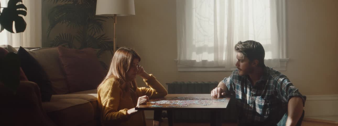 A man and a woman sitting on the floor at a table playing with a puzzle with a wall and window in the background