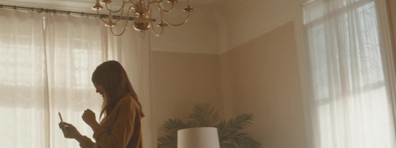 Woman with brown hair and a yellow pullover makes a video call on smartphone and walks up and down in the living room at home