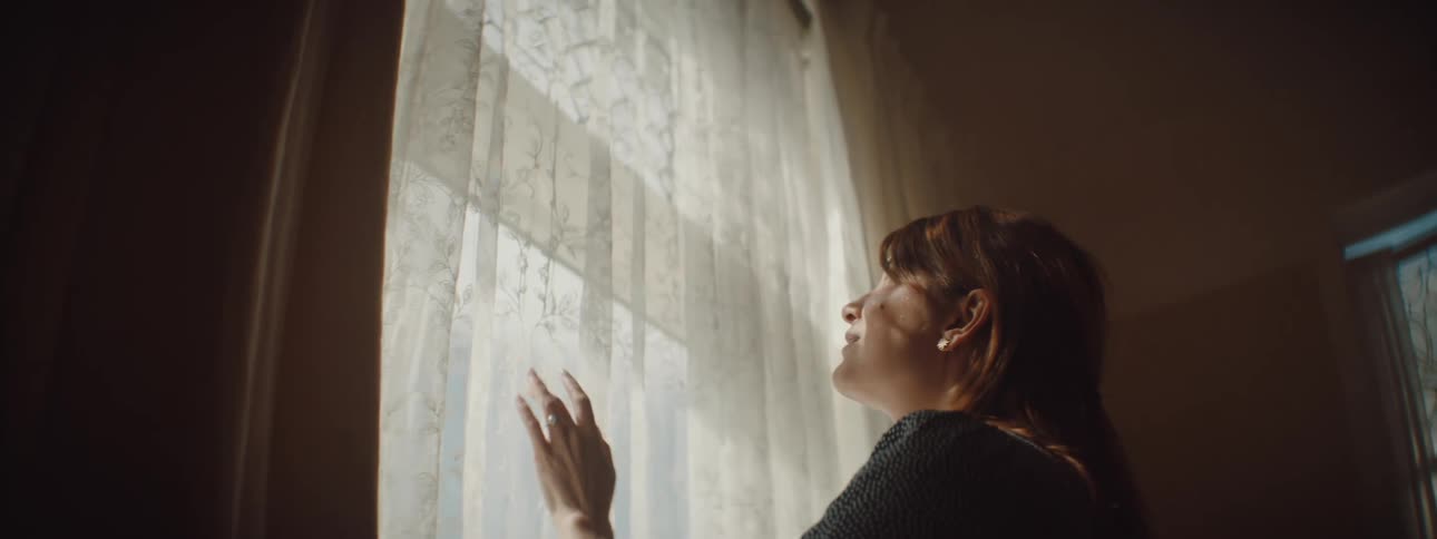 Woman with a blue spotted dress touches the curtains and looks out the window