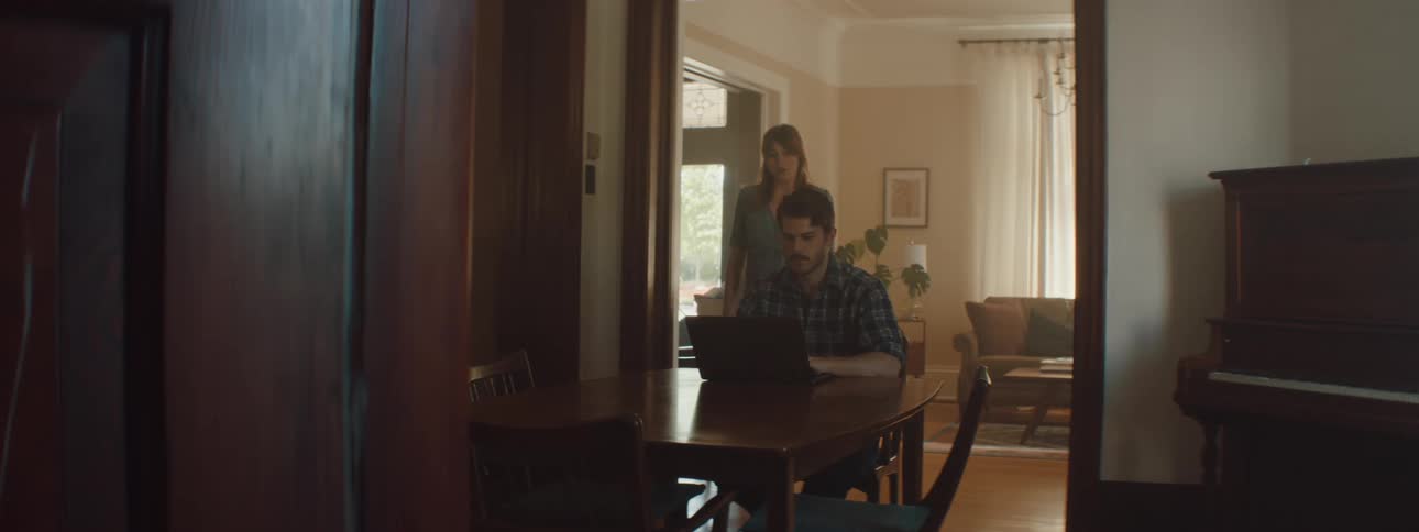 Woman with brown hair and a dress approaches man sitting at the table and using a laptop next to the piano