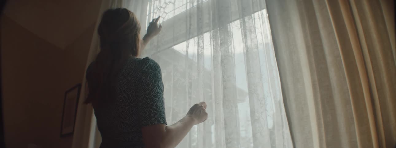 Woman with brown hair and a dress touches the curtains and looks out the window