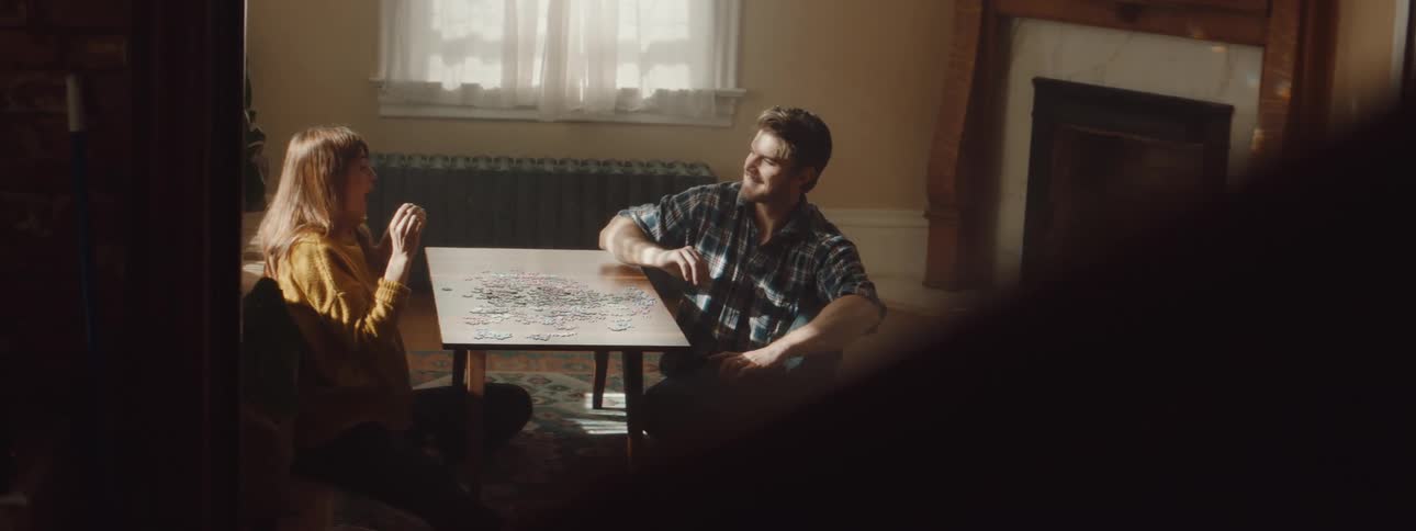 A couple laugh and start throwing puzzle pieces at each other while sitting at a table with a jigsaw puzzle